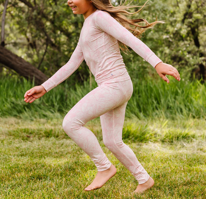 A girl wearing a light pink long-sleeve top and matching leggings runs barefoot across a grassy outdoor area with trees in the background.