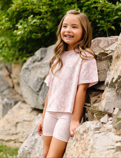 A young girl with long light brown hair stands outdoors against a rocky wall with green foliage behind her, wearing a light pink short-sleeve t-shirt and matching light pink shorts.