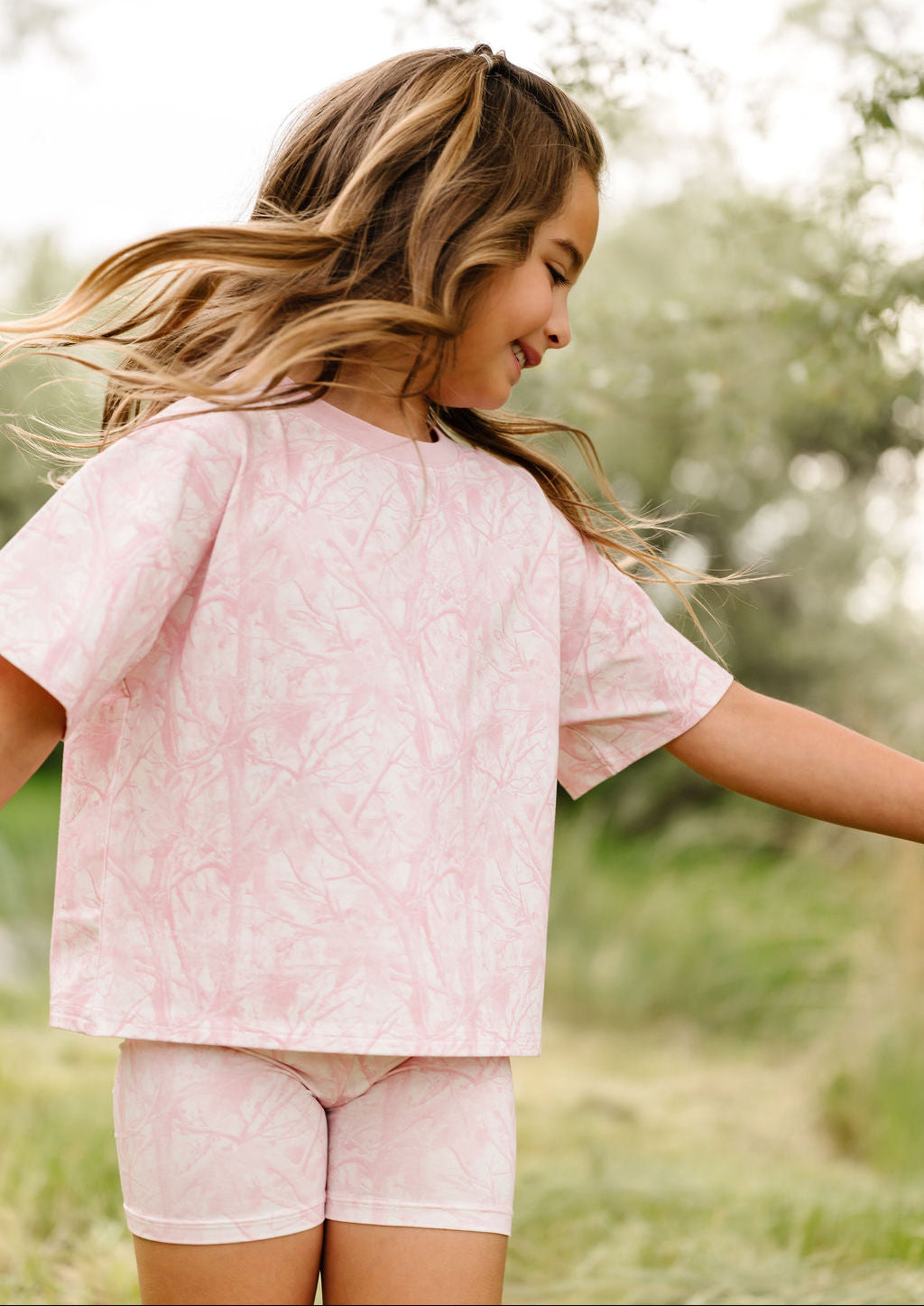 A young girl with long blonde hair is wearing a light pink short-sleeve t-shirt and matching shorts with a tonal leaf pattern, set against a soft-focus outdoor background of green grass and trees.
