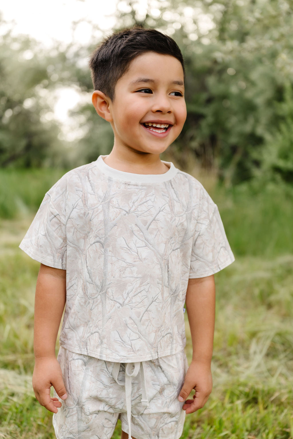Child wearing a light-colored tree camo outfit, standing in a grassy outdoor setting.