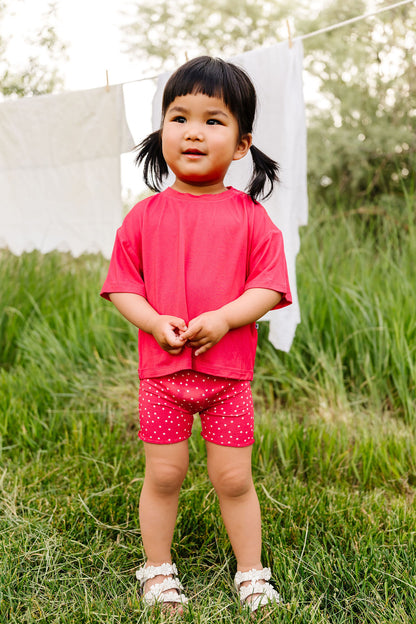 Child wearing a red shirt and berry pink shorts with white polka dots standing in a grassy area.