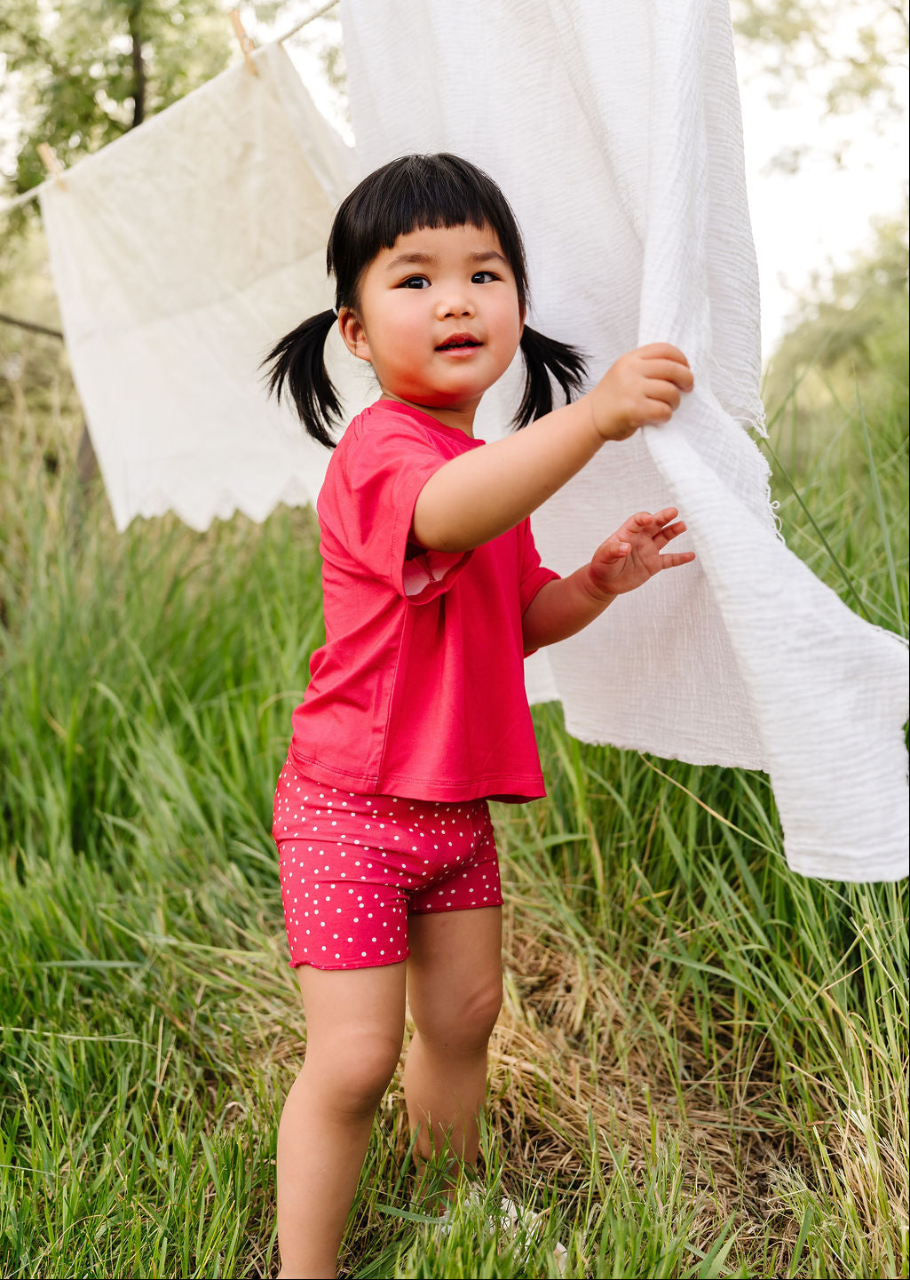 Child in a red shirt and shorts standing in a grassy field with white fabric hanging in the background.