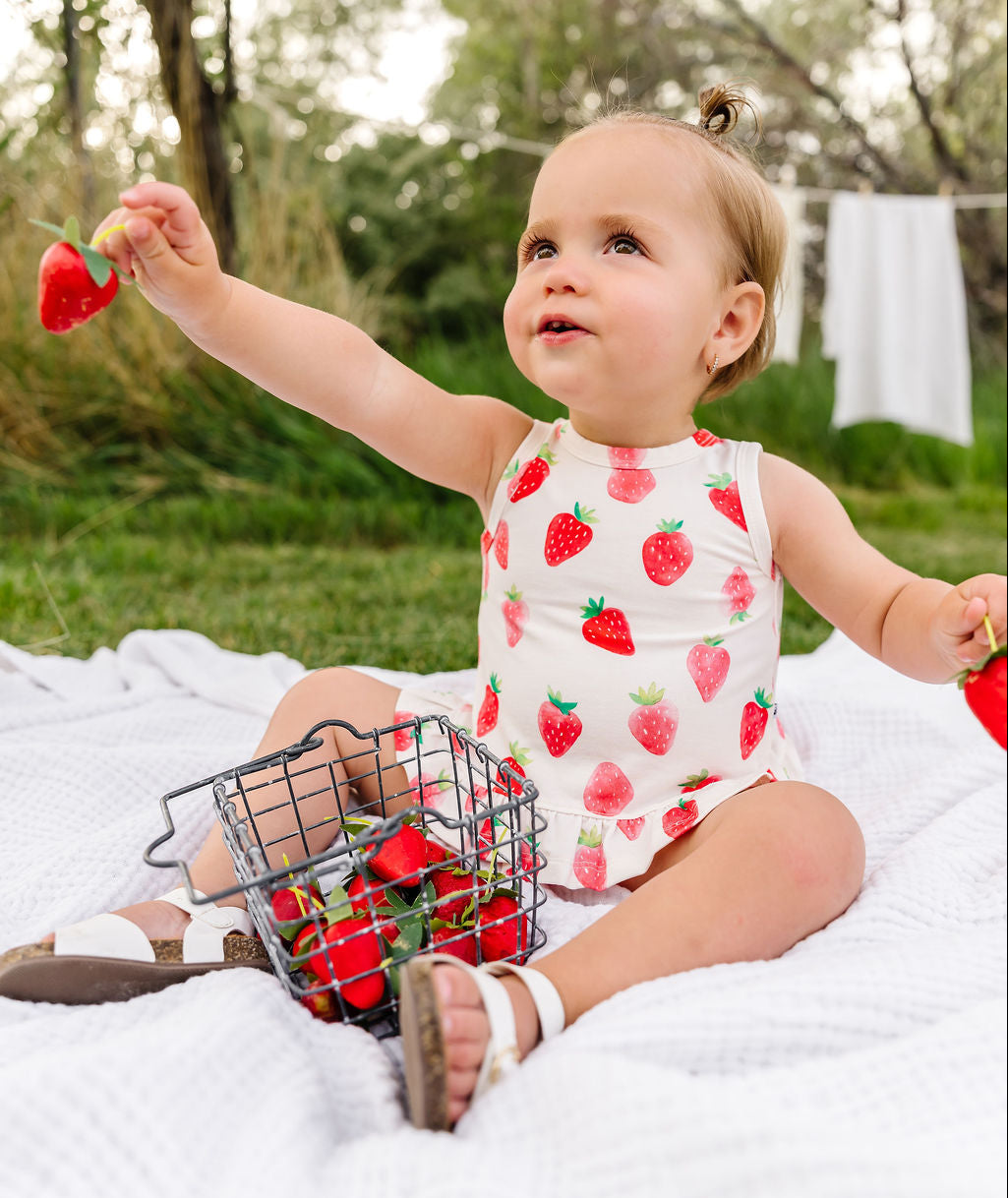 Child in a strawberry-patterned outfit sitting on a blanket outdoors with a basket of red toys.