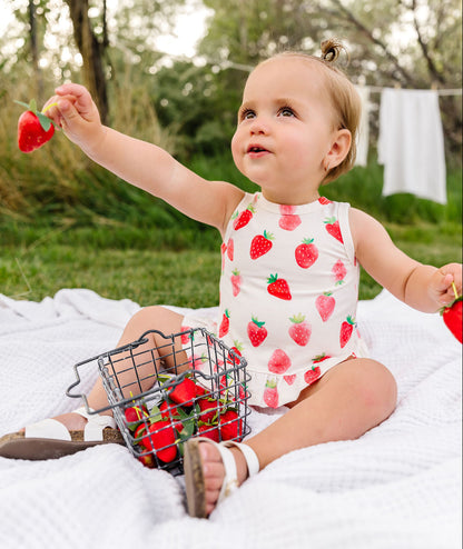 Child in a strawberry-patterned outfit sitting on a blanket outdoors with a basket of red toys.