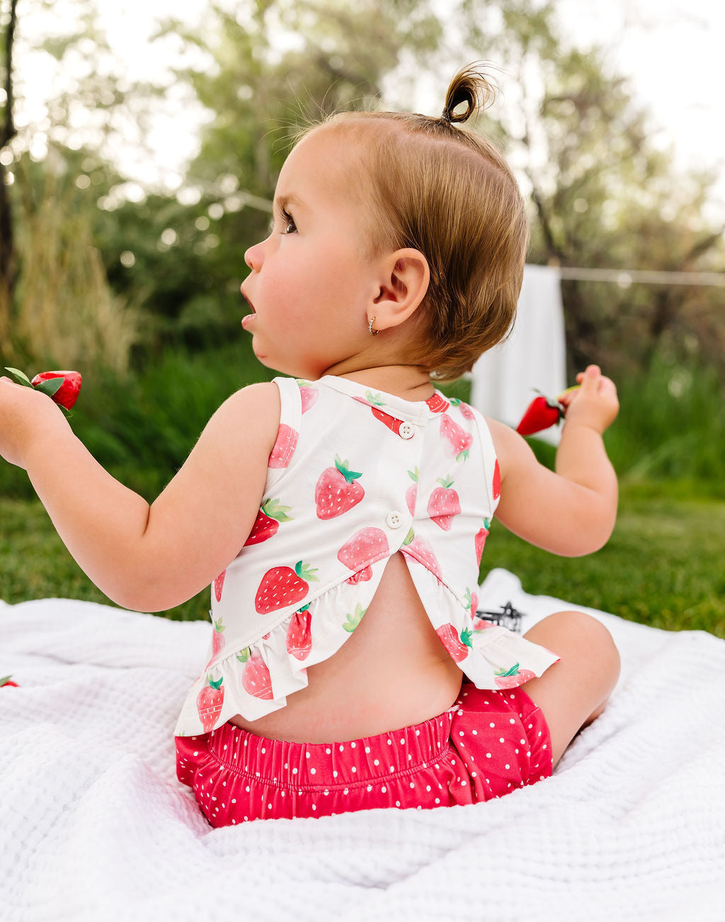 Baby sitting on a blanket outdoors wearing a strawberry-patterned outfit.