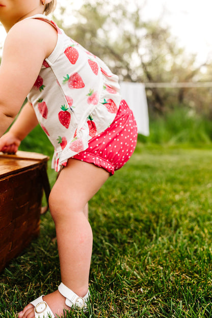 Child in a strawberry-patterned top and red polka dot shorts standing on grass.