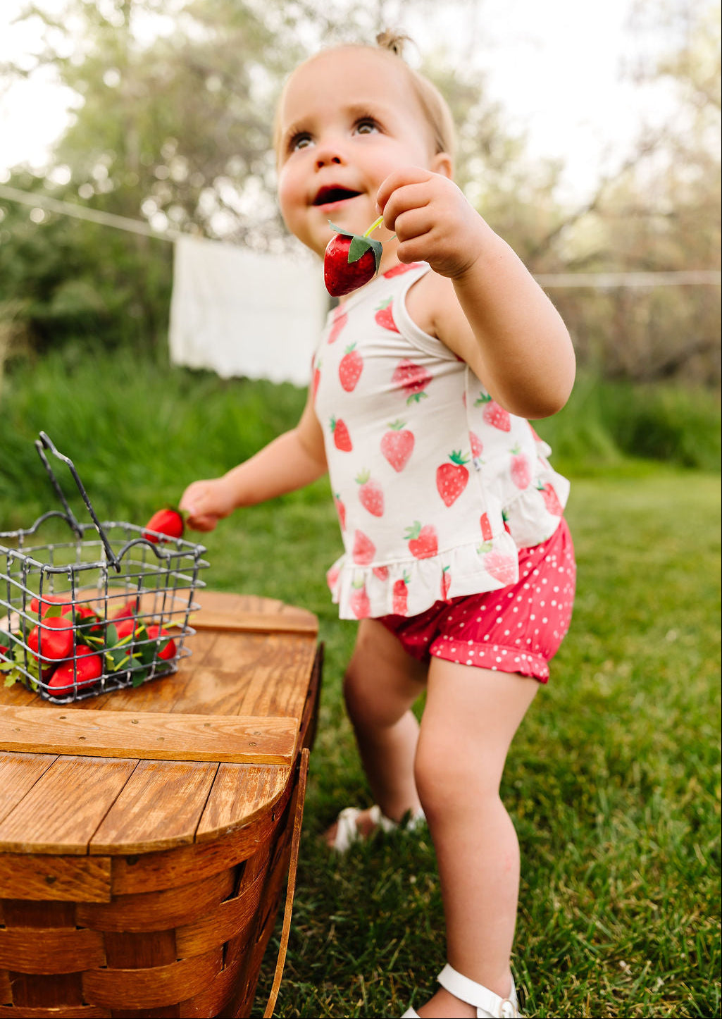 Child holding a strawberry outdoors near a wooden table with a basket of strawberries.