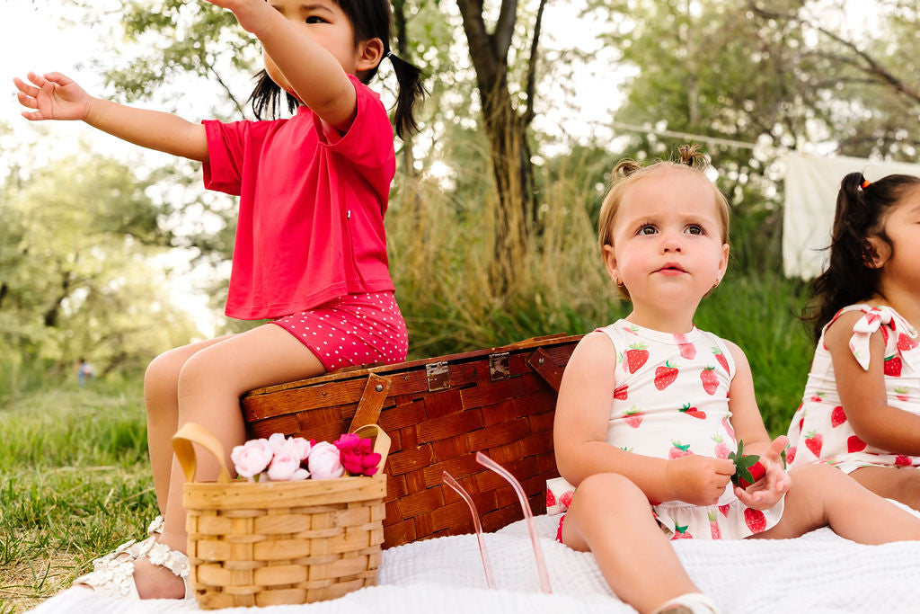Three children sitting on a picnic blanket with a basket and picnic items in a park.