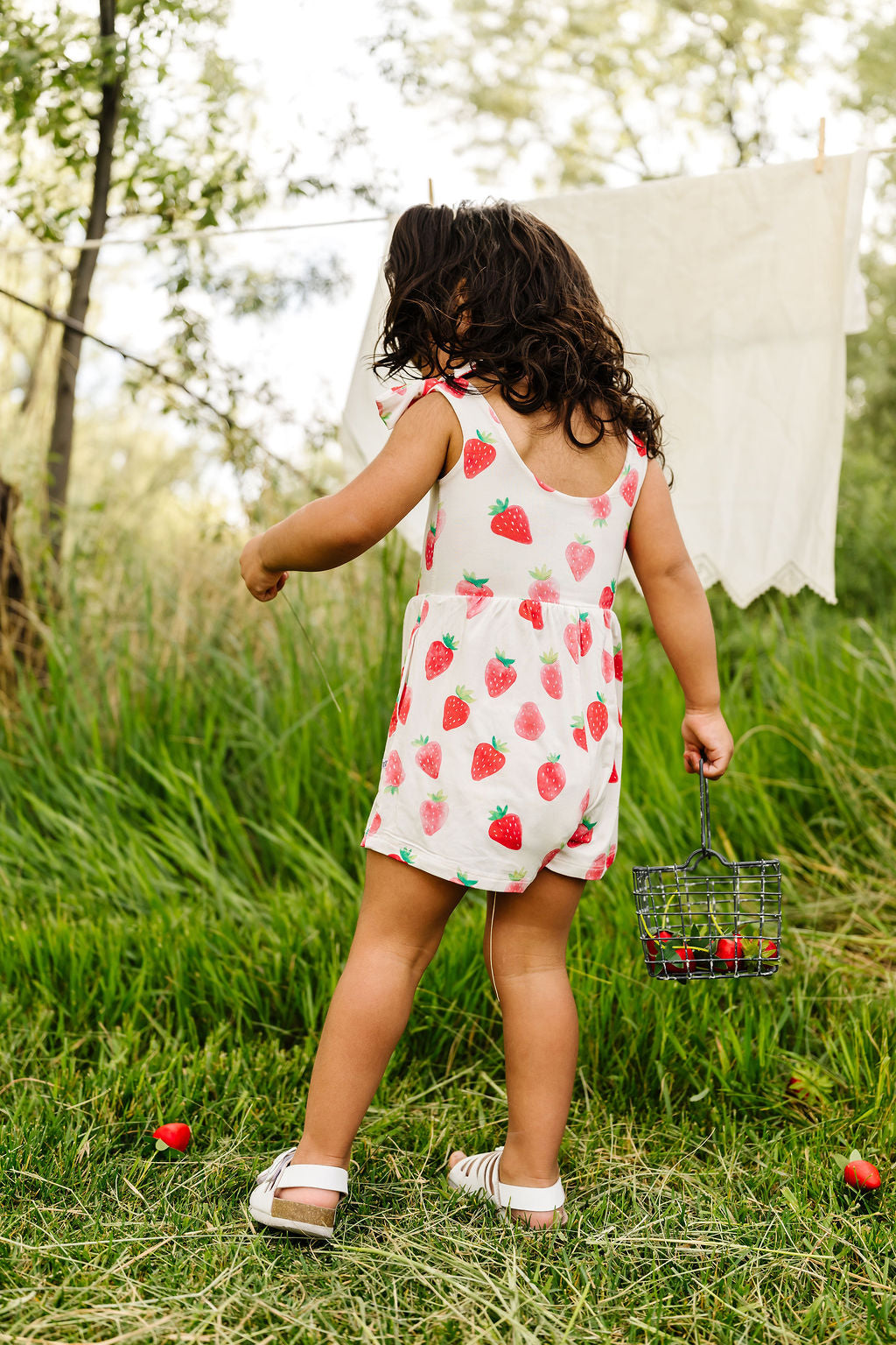 Child in a strawberry romper standing in a grassy field with a laundry basket and scattered strawberries.
