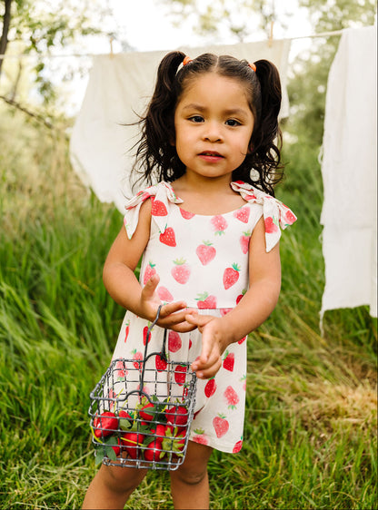 Young girl in a strawberry romper holding a basket of berries in a natural setting.