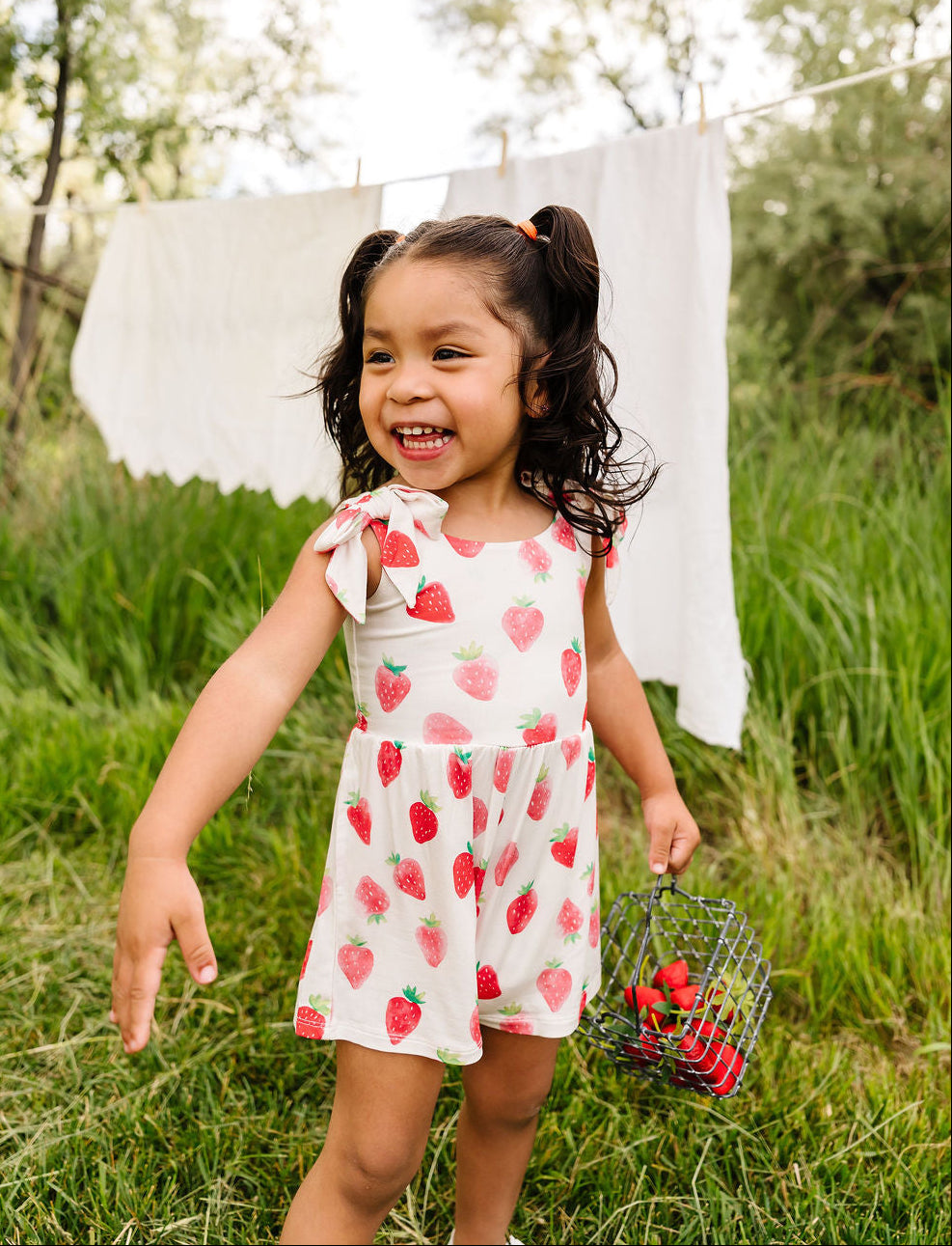 Young girl in a strawberry outfit holding a basket of strawberries in a grassy outdoor setting.