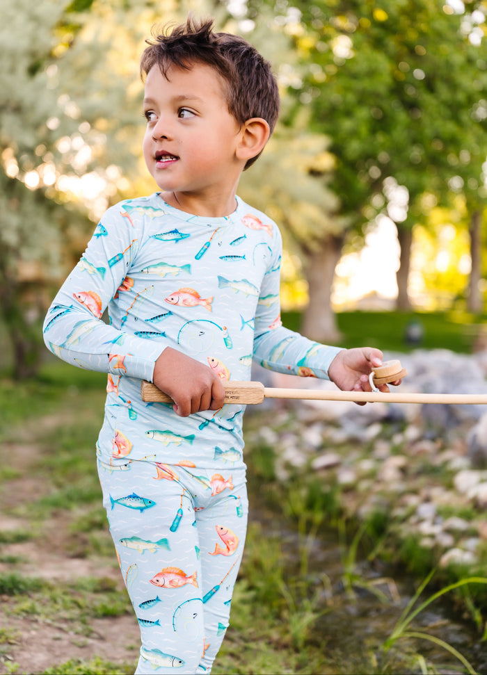 Little boy in a light blue bamboo pajama set with fish pattern holding a fishing rod outdoors.