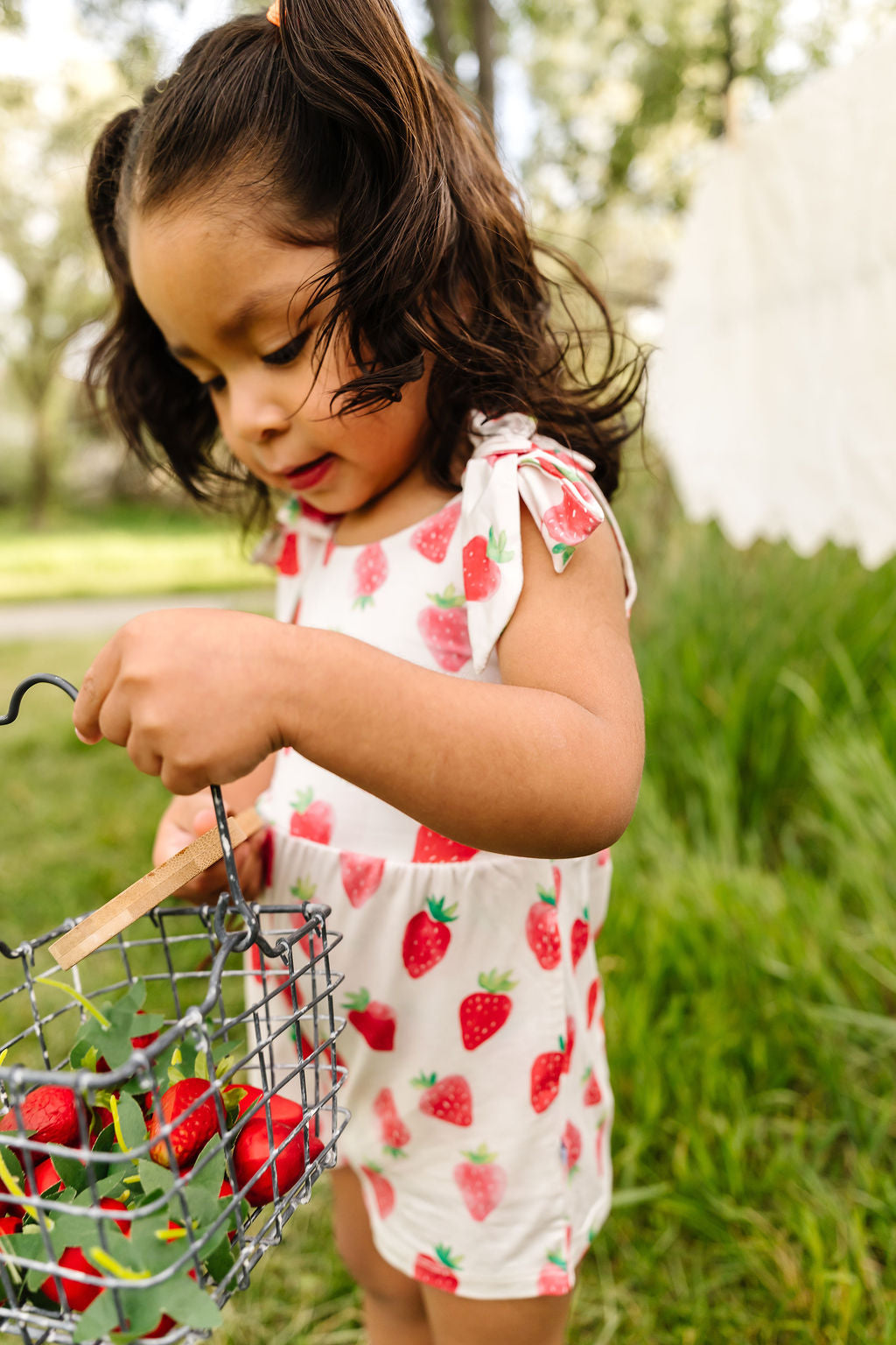 Young girl in a strawberry outfit holding a basket of strawberries outdoors.