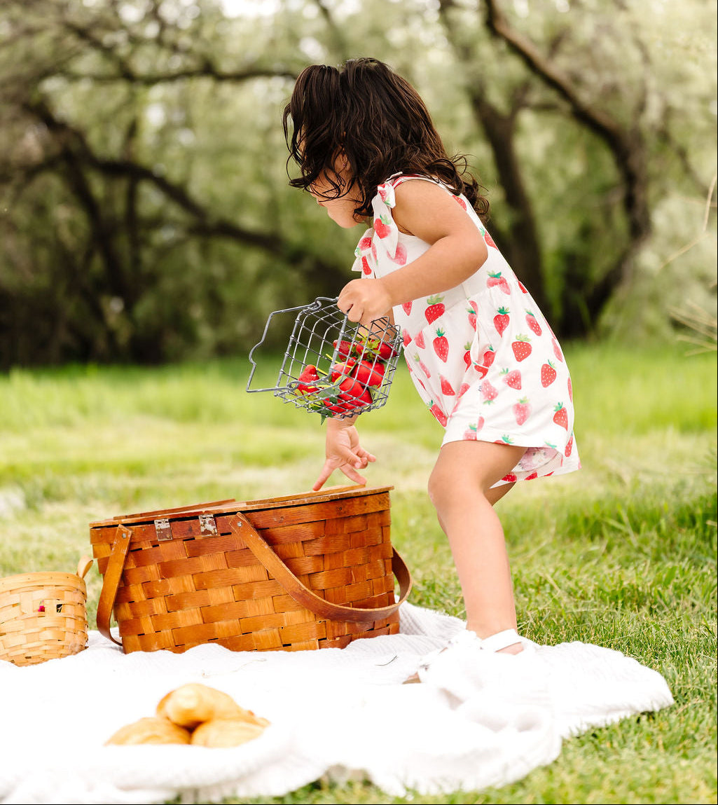 Child in a strawberry dress picking berries near a picnic basket in a park.