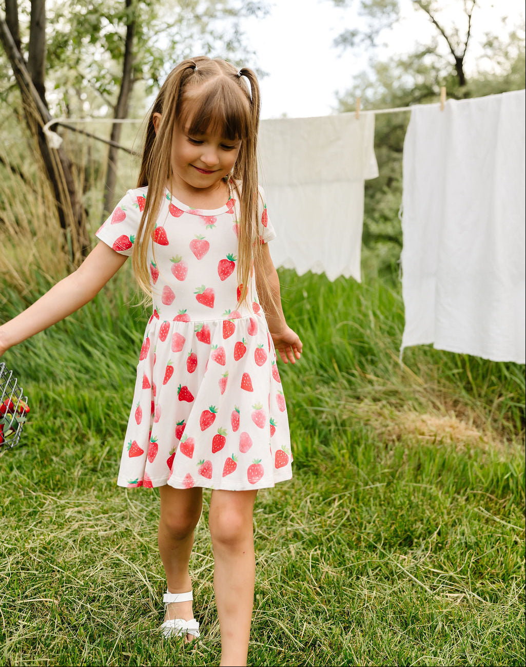 Young girl in a strawberry dress standing in a grassy area with trees and laundry in the background.