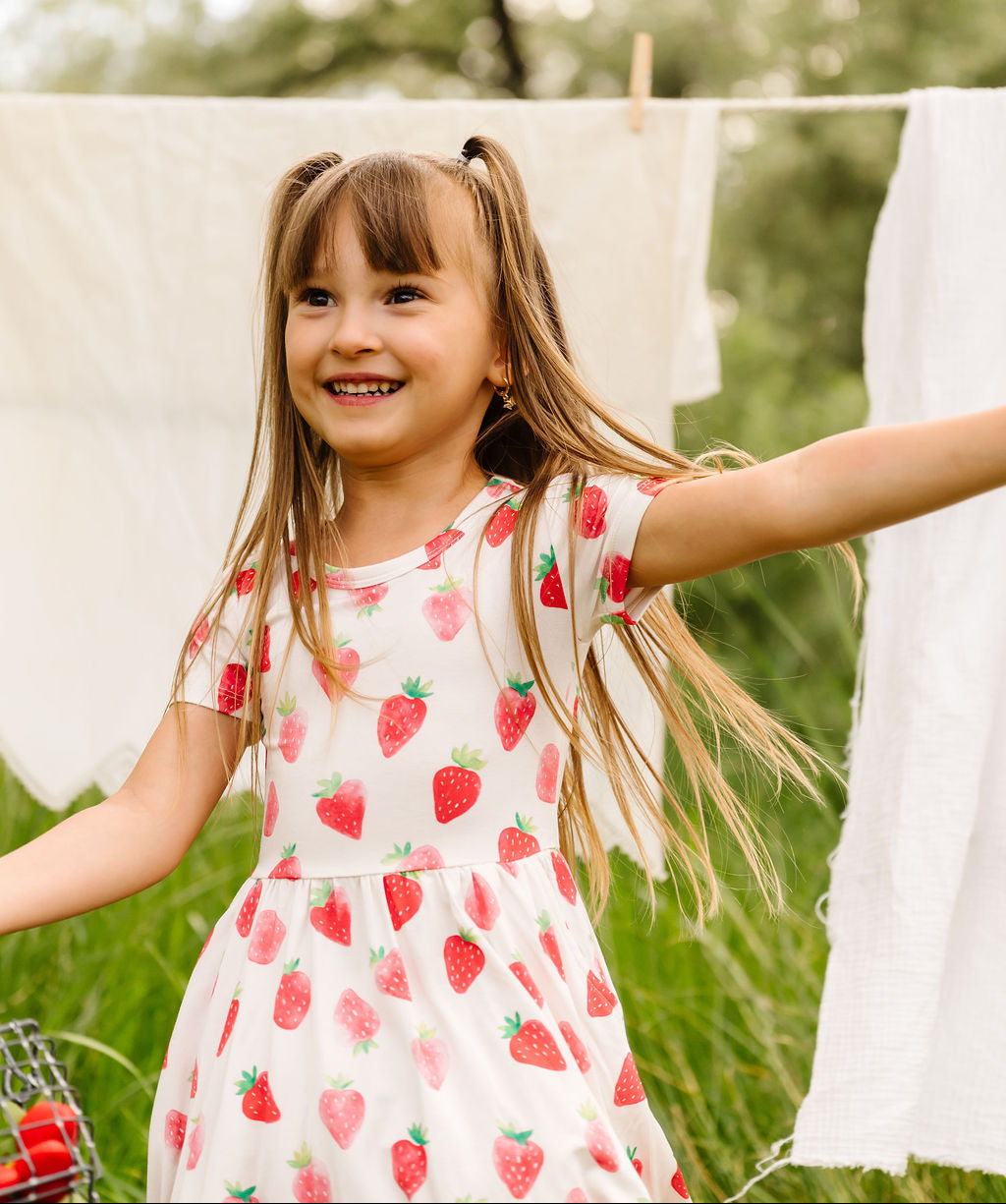 Young girl in a strawberry dress standing outdoors with greenery in the background