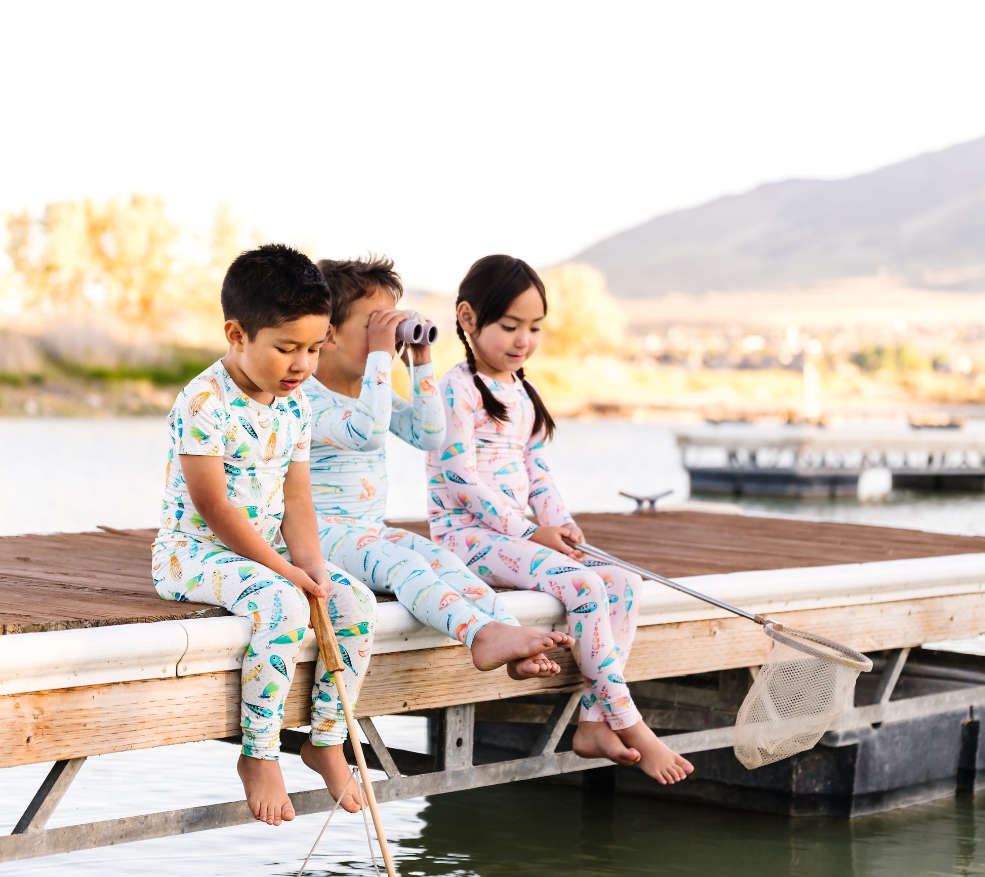 Three children in pajamas sitting on a dock by a body of water at dusk.