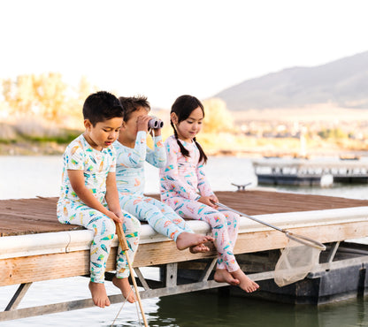 Three children in pajamas sitting on a dock by a body of water at dusk.