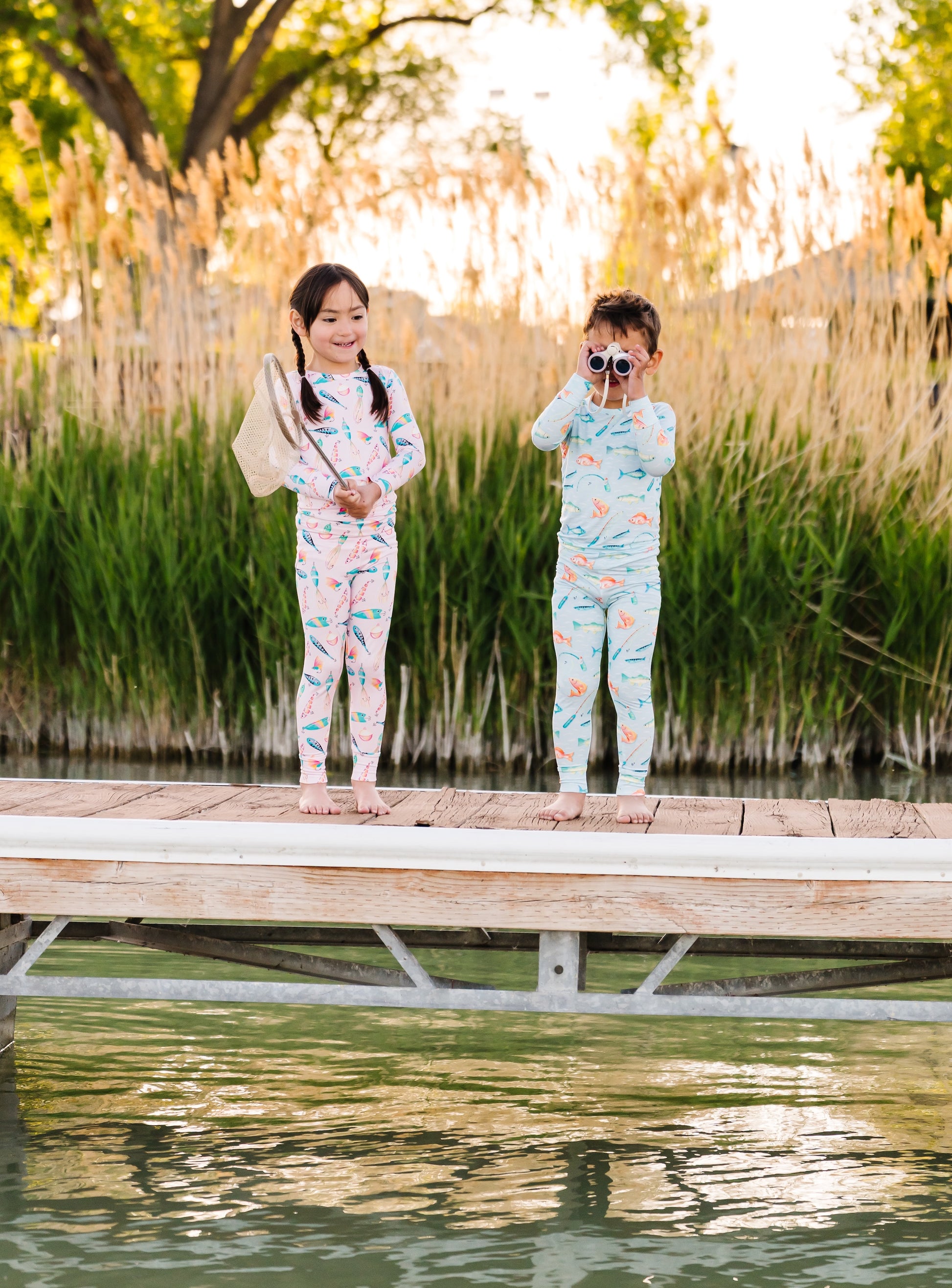 Two children in pajamas standing on a dock by a body of water with reeds in the background.