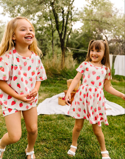 Two young girls in matching strawberry print outfits standing outdoors on a grassy area with trees in the background.