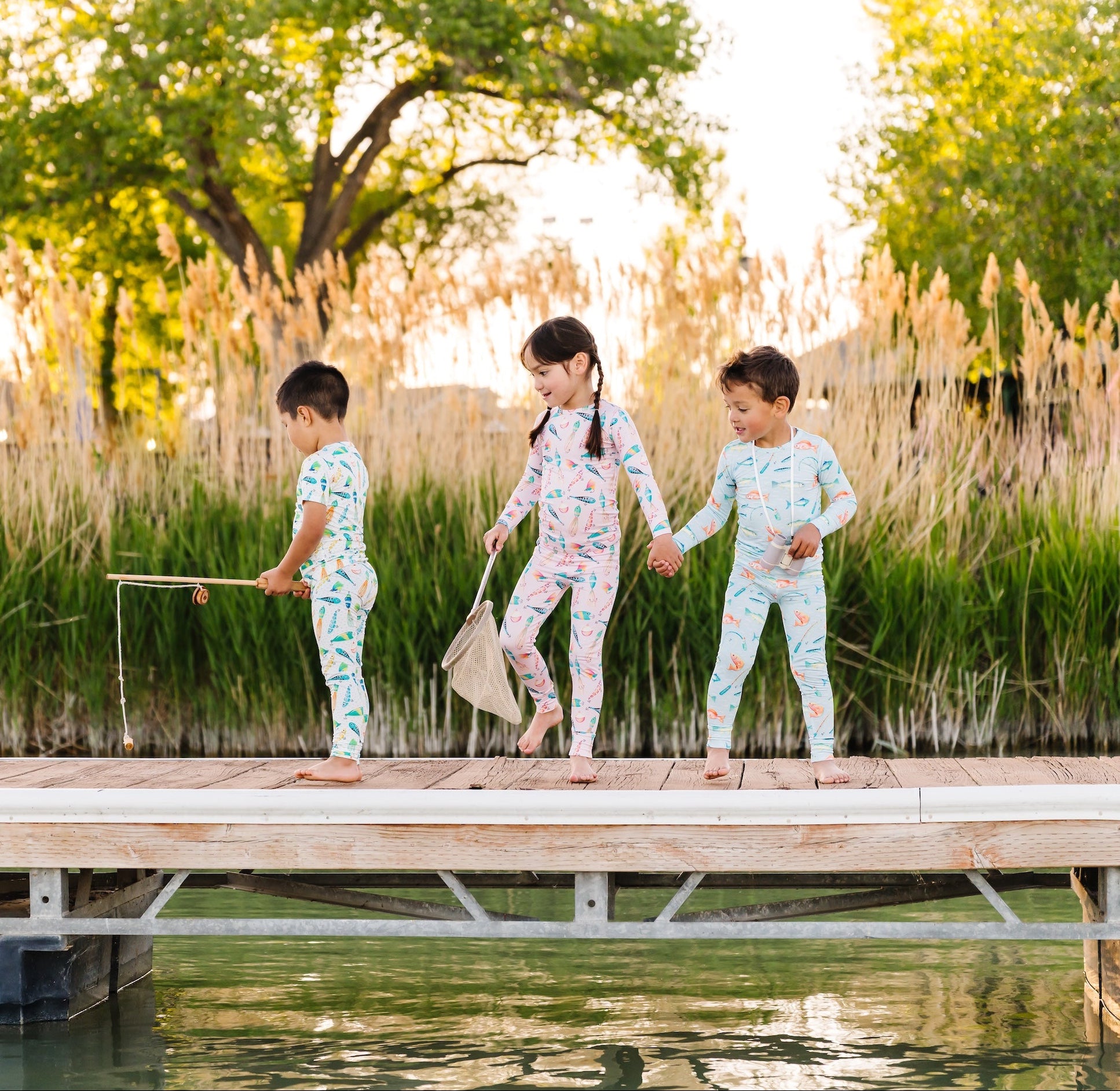 Three children in matching pajamas standing on a dock by a body of water with trees and grass in the background.