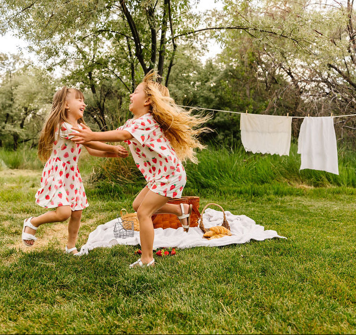 Two girls in strawberry outfits dancing outdoors with a picnic basket and blanket