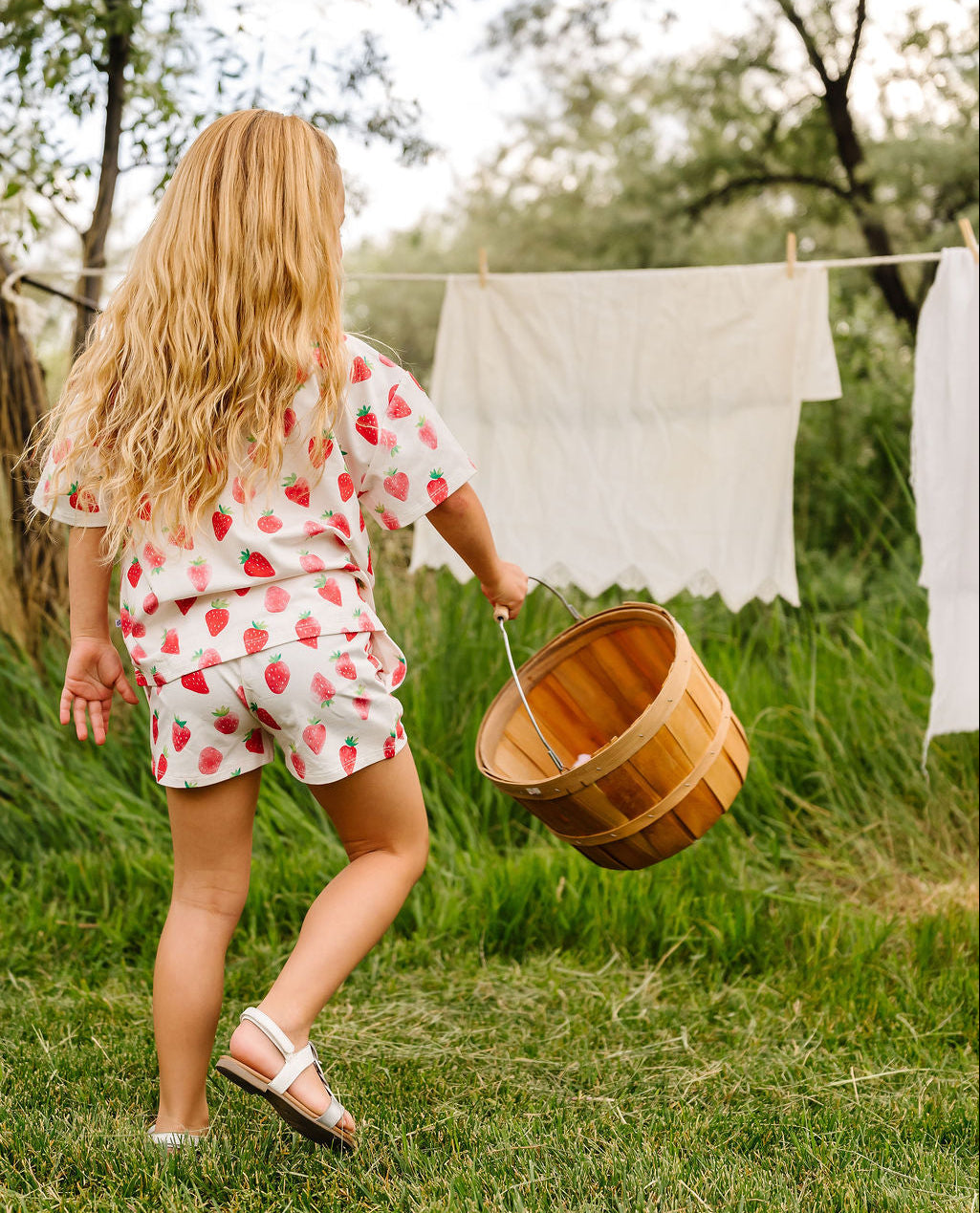 Child in a red and white outfit carrying a wooden basket in a grassy outdoor setting with clothes hanging on a line.