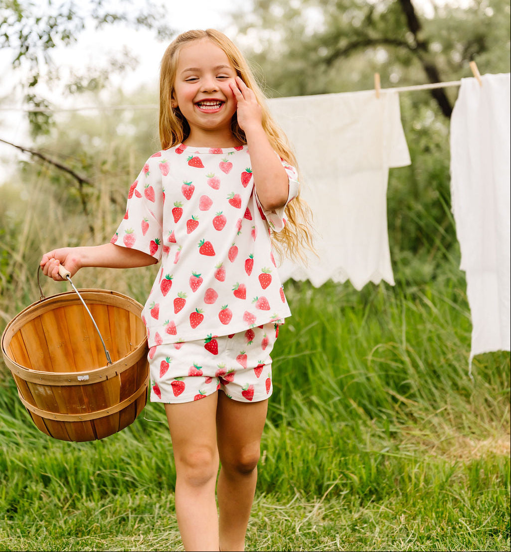 Young girl in a strawberry print outfit holding a wooden basket outdoors.