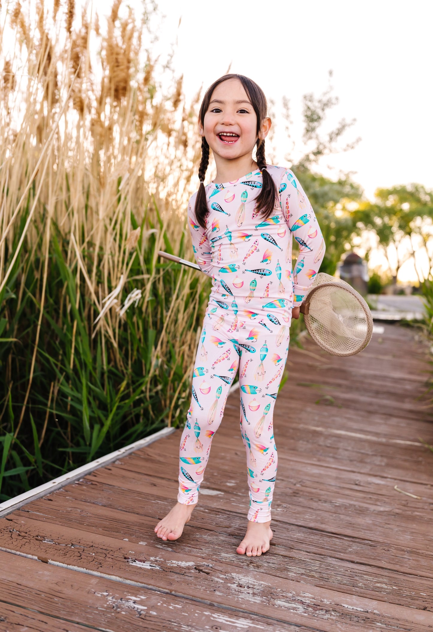 Child wearing a fishing-patterned pajama set standing on a wooden dock with tall grasses in the background.