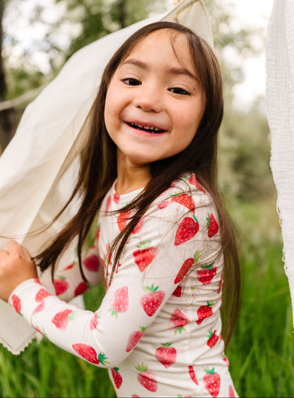 Young girl in a strawberry-patterned pajama set standing outdoors with greenery in the background