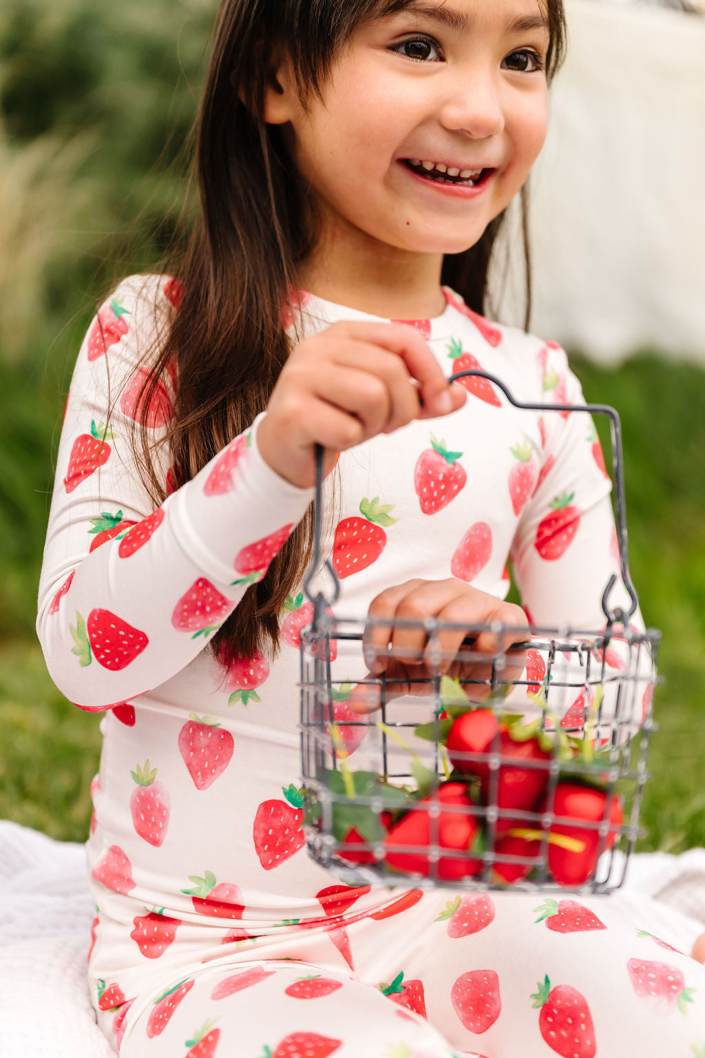 Child wearing strawberry-patterned pajamas holding a basket of strawberries outdoors.