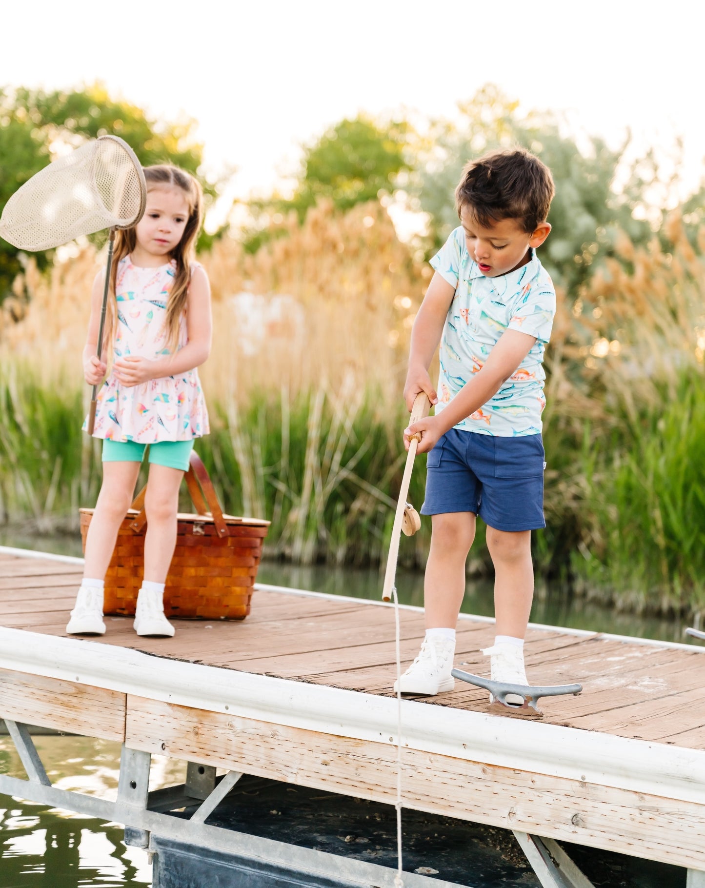 Two children on a wooden dock by a body of water, with one holding a fishing net and the other standing.