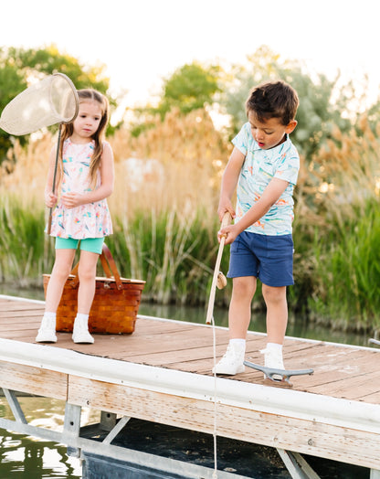Two children on a wooden dock by a body of water, with one holding a fishing net and the other standing.