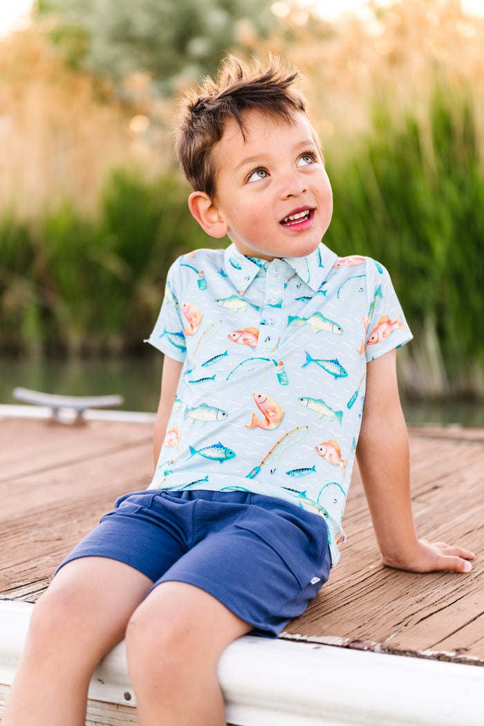 Young boy sitting on a dock by a body of water, wearing a bamboo fishing polo and blue shorts.