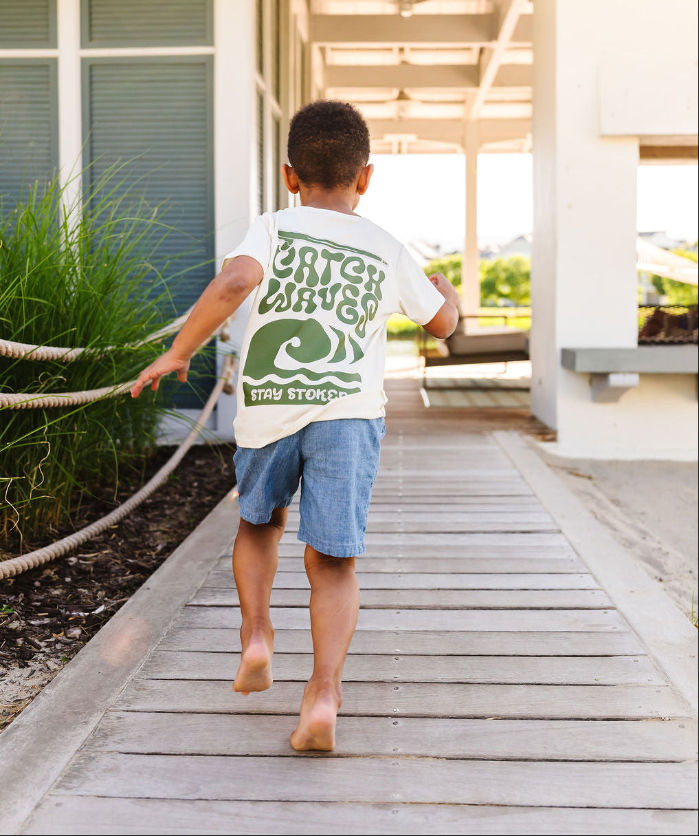 Child walking on a wooden deck wearing a white t-shirt with green text and graphics.