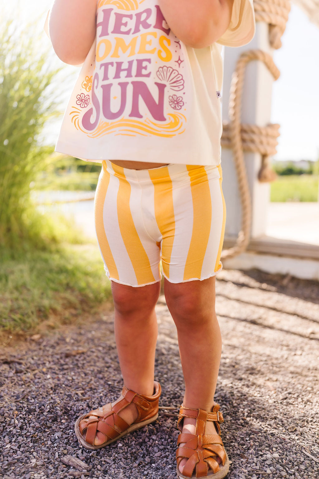 Child wearing a sleeveless top with 'Here Comes the Sun' text and yellow striped shorts outdoors.