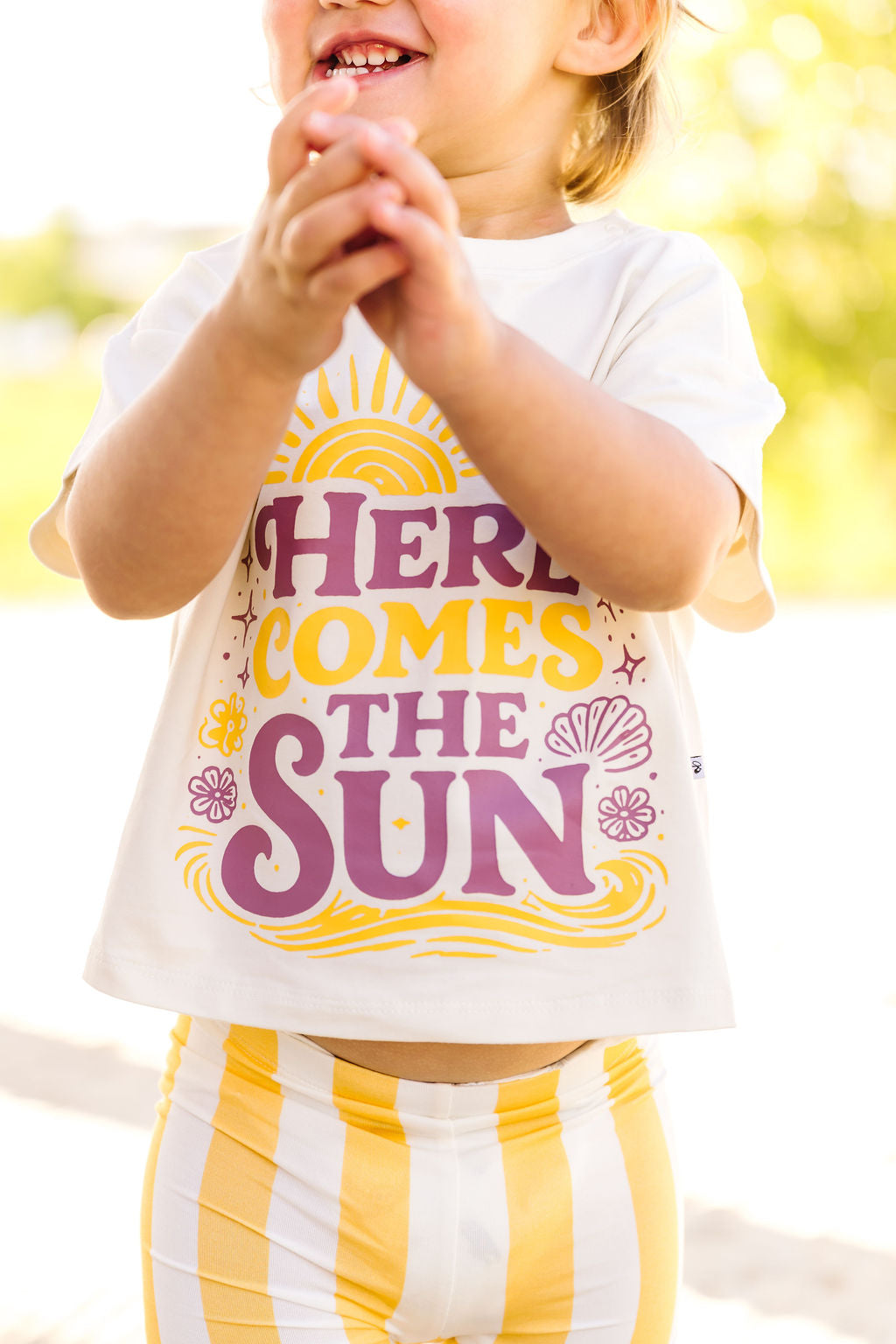 Child wearing a shirt with 'Here Comes the Sun' text and sun graphic outdoors.