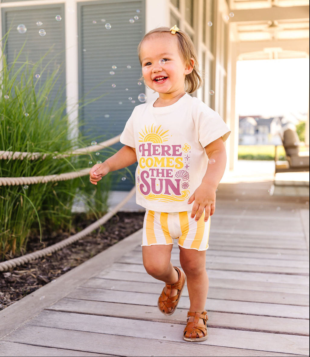 Child wearing a white t-shirt with colorful text and yellow shorts, walking on a wooden deck.