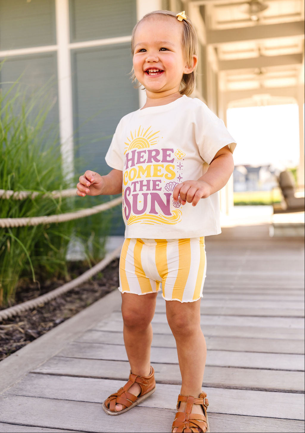 Child wearing a white t-shirt with colorful text and yellow shorts standing on a wooden deck.