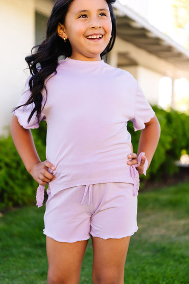 Young girl in a light purple outfit standing outdoors with greenery in the background