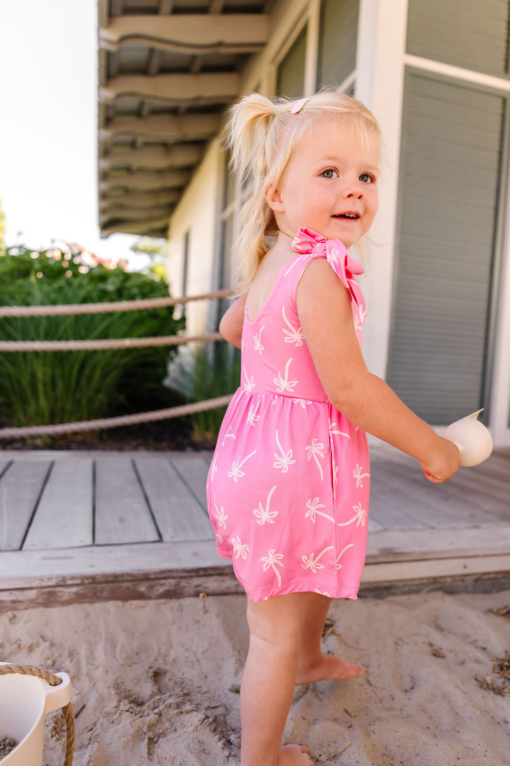 Young girl in a pink romper with white patterns standing on a sandy area.
