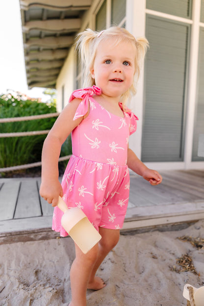 Young girl in a pink romper with white patterns standing on a sandy surface.