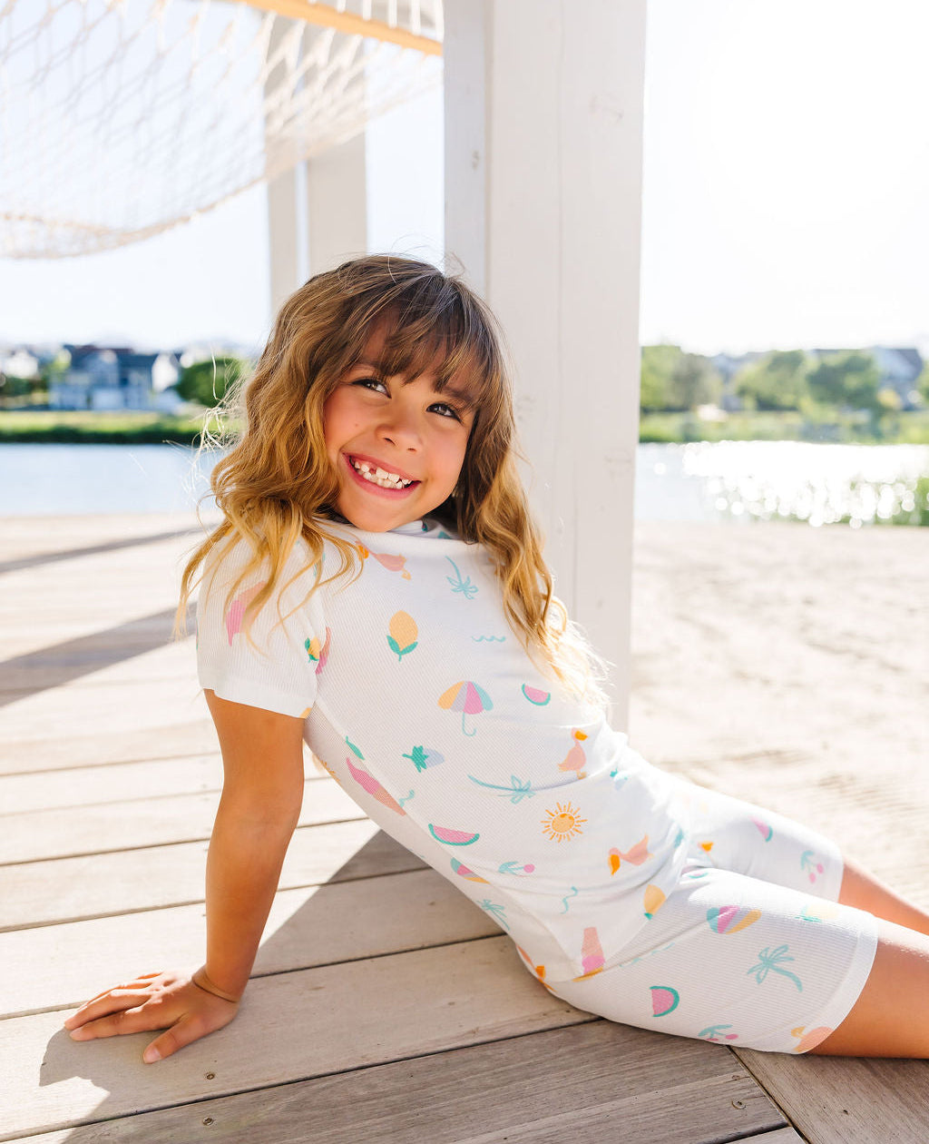 Child wearing a white outfit with colorful patterns sitting on a wooden deck by a waterfront.