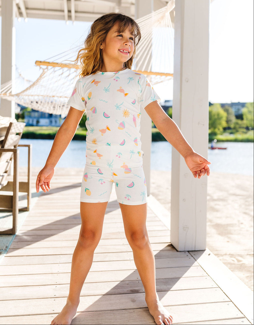Child wearing a pajama set with colorful patterns on a wooden deck by a waterfront.