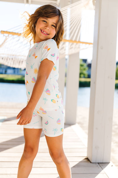 Child wearing a colorful pj set with a pattern of palm trees, umbrellas, and lemons, standing on a wooden deck by a body of water.