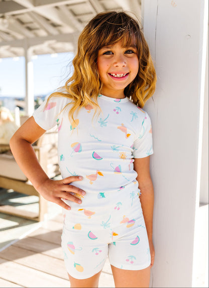 Young girl wearing a white t-shirt and white shorts with colorful patterns, standing on a wooden deck.