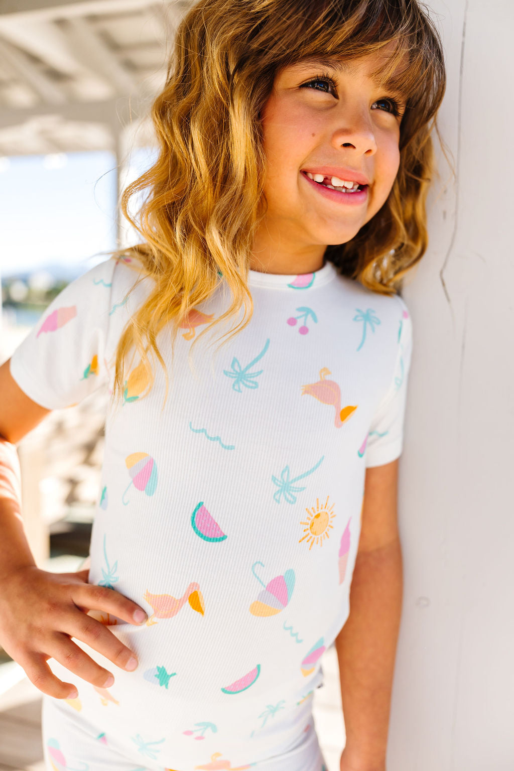 Child wearing a white pj shirt with colorful patterns, standing against a white wall.