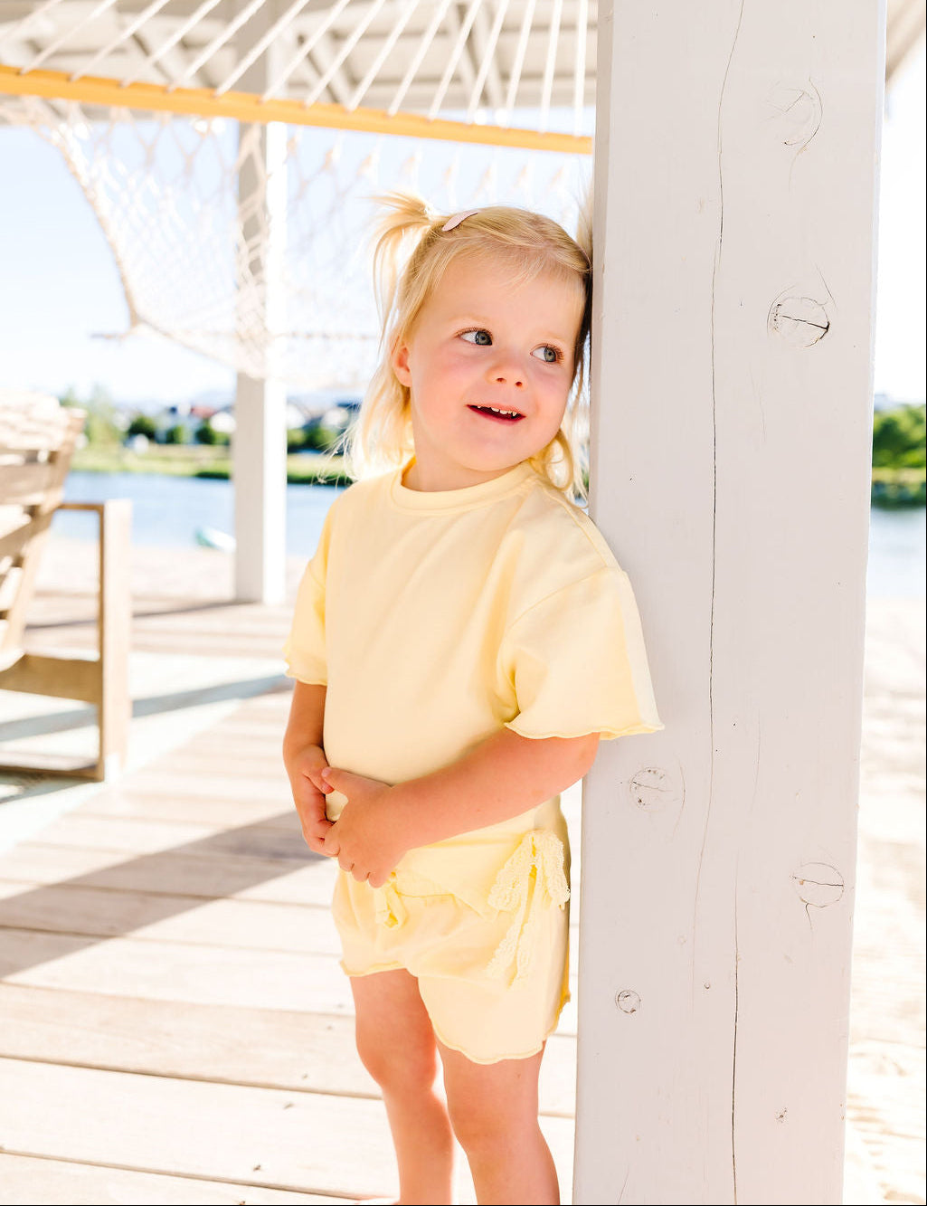 Young girl in a yellow outfit standing on a wooden deck with a hammock in the background.