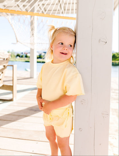 Young girl in a yellow outfit standing on a wooden deck with a hammock in the background.