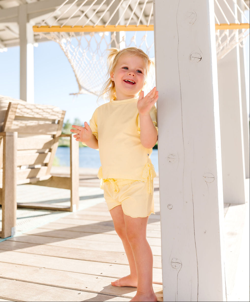 Child in a yellow outfit standing on a wooden deck with a hammock in the background.